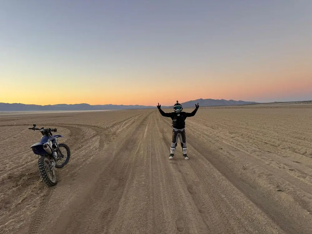 A rider with arms raised on a desert road at sunset, dirt bike parked nearby